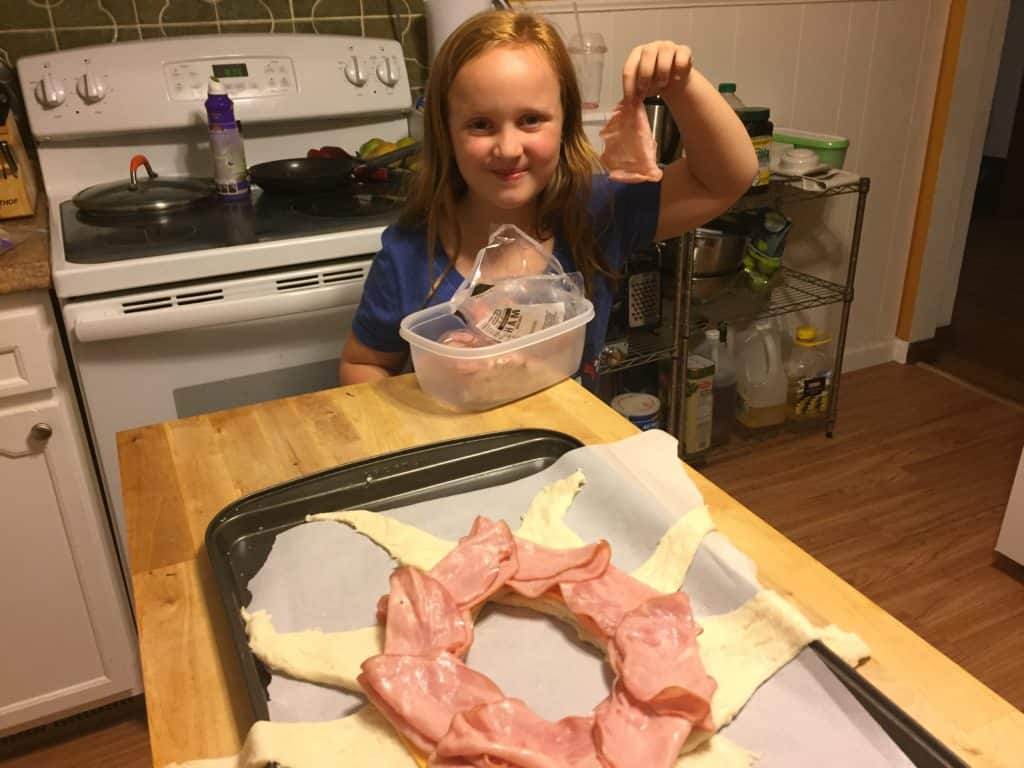 daughter standing in kitchen holding a slice of ham helping assemble ham and cheese crescent ring