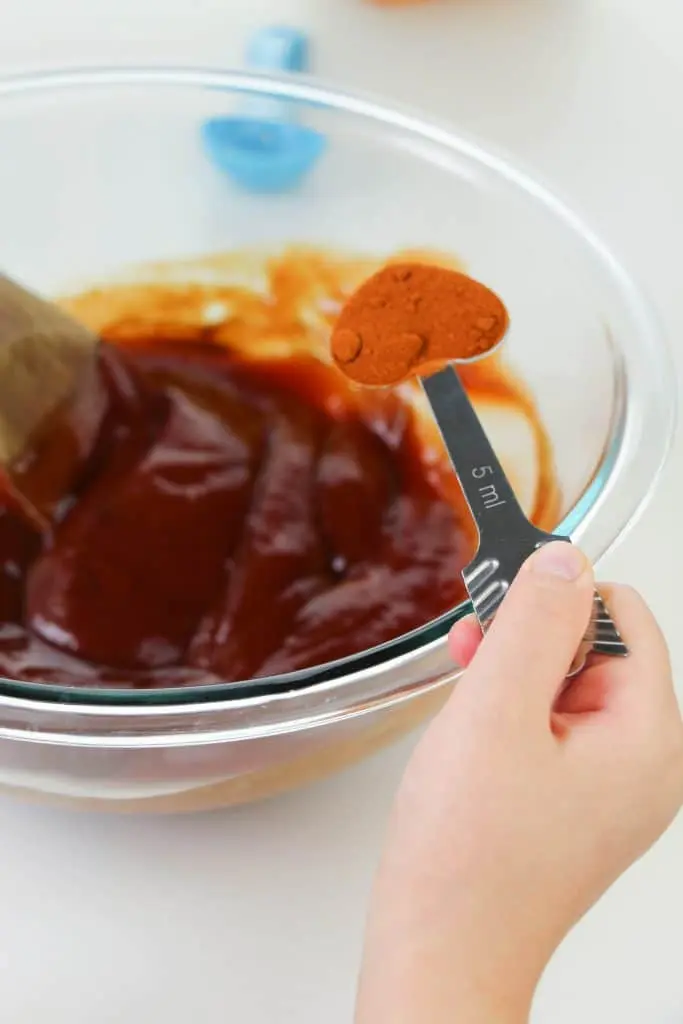 homemade bbq sauce ingredients in clear glass bowl with child's hand adding tablespoon of paprika