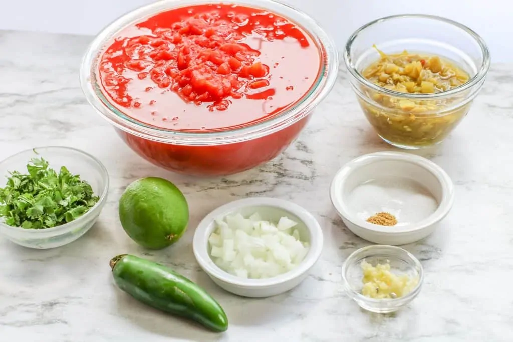 canned tomatoes canned green chilis cilantro jalapeno lime onion and spices in glass bowls on marble countertop