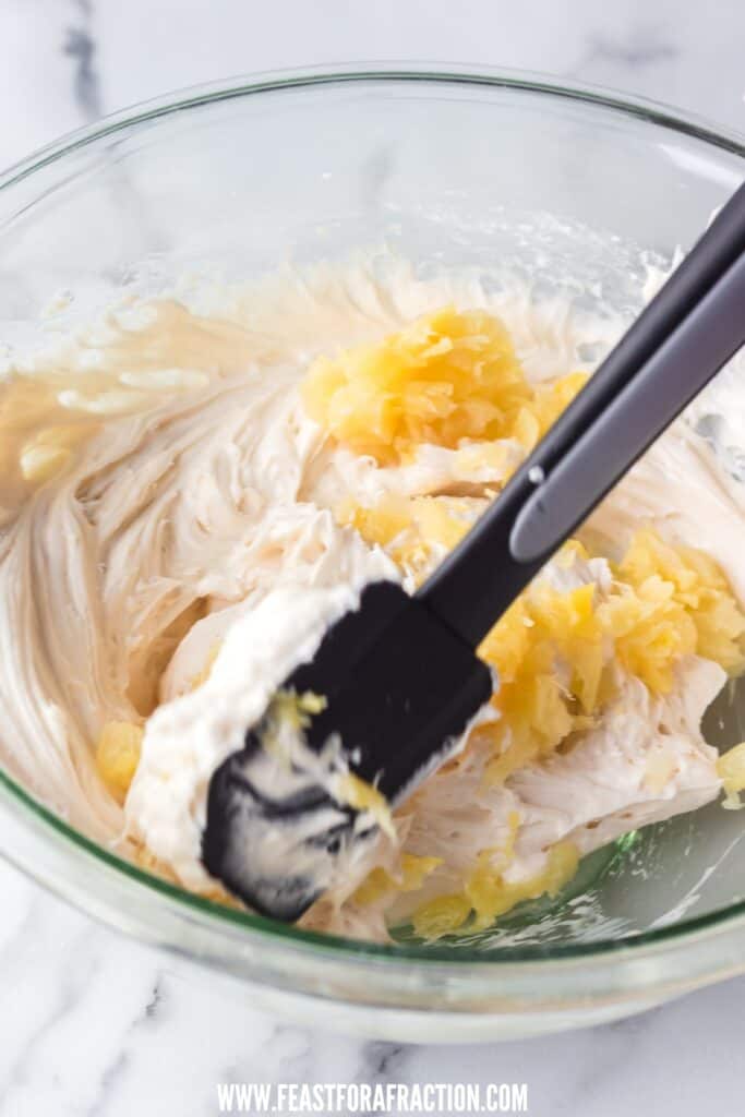 A glass bowl containing cream cheese frosting being mixed with crushed pineapple, using a black spatula, on a marble surface, intended for whoopie pies.