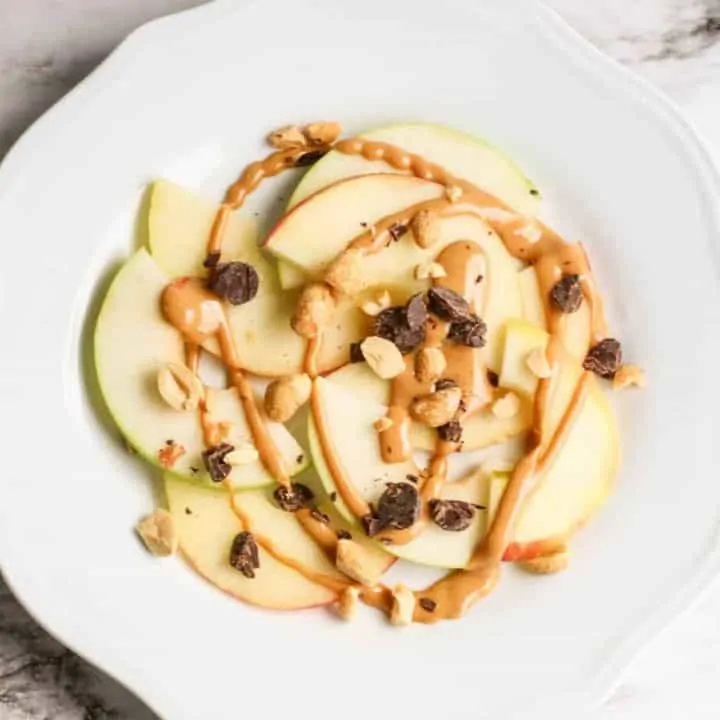 sliced apples on white plate topped with nut butter and chopped nuts and chocolate for apple nachos
