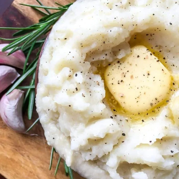 overhead view of mashed potatoes in a brown bowl with butter and garlic and rosemary on wooded board