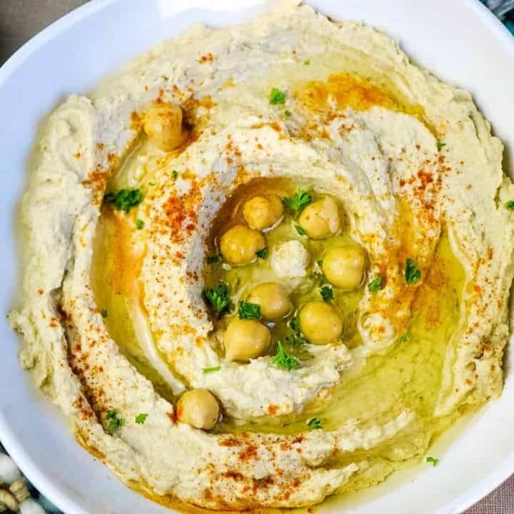 square image of hummus in white bowl with whole chickpeas and chopped parsley