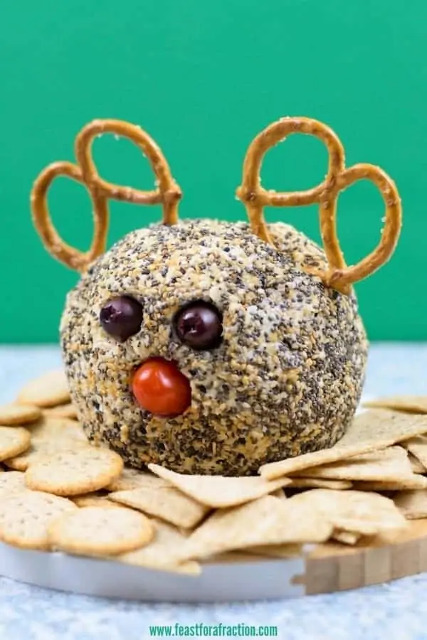 cheese ball coated in everything bagel seasoning with pretzels, black olives and tomato to look like reindeer