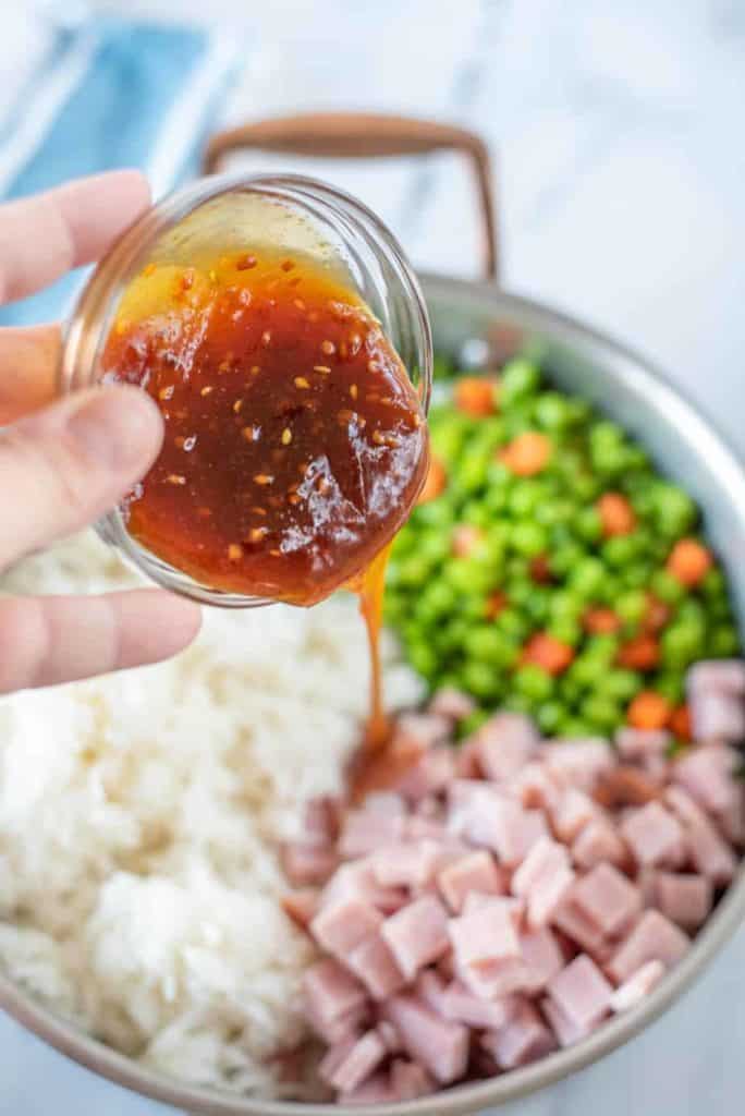 teriyaki sauce being poured into pan with white rice, diced ham and peas and carrots