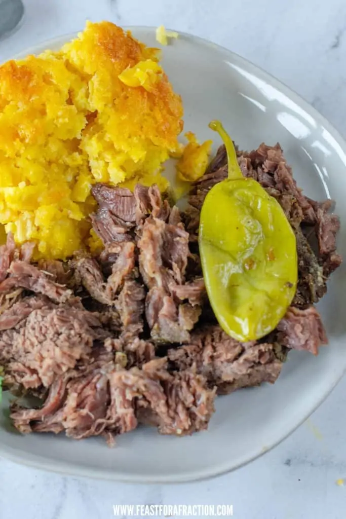 cooked crockpot mississippi pot roast on white plate with corn casserole and pepperoncini pepper