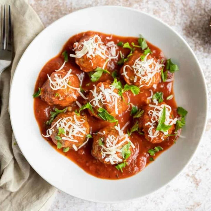 overhead view of baked meatballs in white bowl with tomato sauce, shredded parmesan cheese and chopped parsley