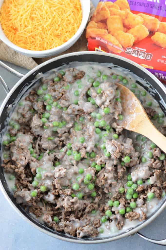cooked ground beef, cream of mushroom soup and frozen peas in pan being mixed