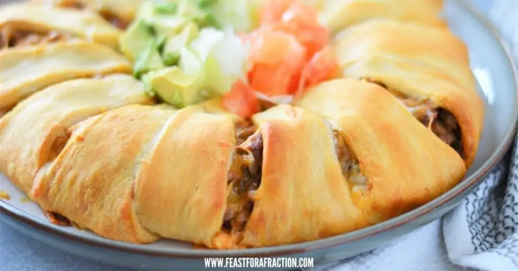 horizontal image of baked taco crescent ring on gray platter garnished with lettuce, tomato and avocado
