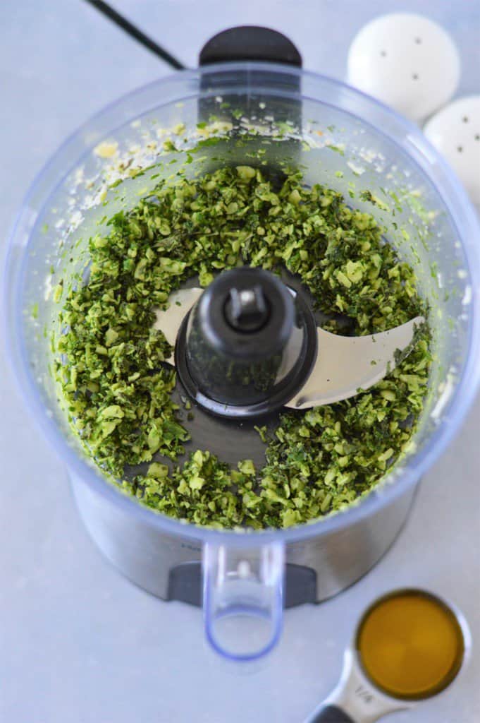 basil, parmesan cheese and garlic blended in food processor with measuring cup of olive oil in background