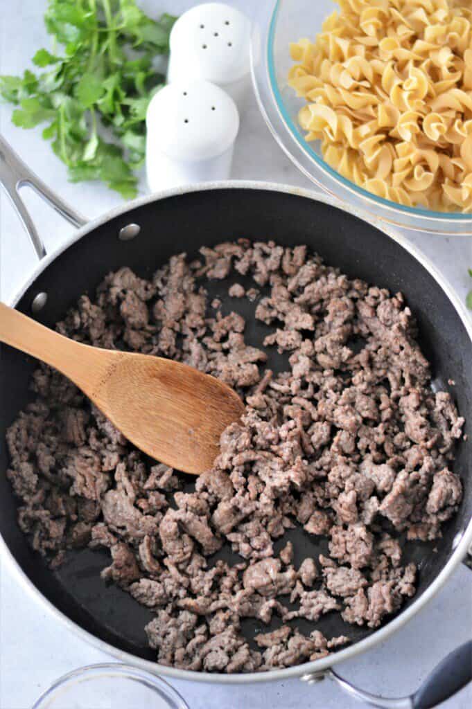 cooked ground beef in pan with wooden spoon and egg noodles in glass bowl in background