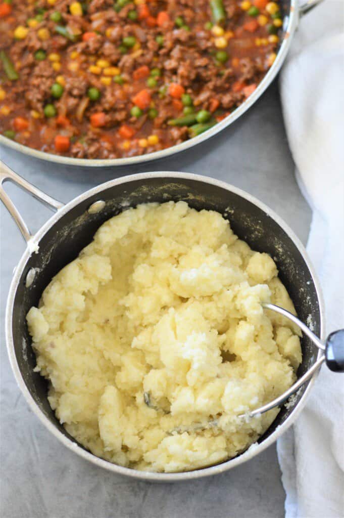 mashed potatoes in pot with potato masher and shepherd's pie filling in background