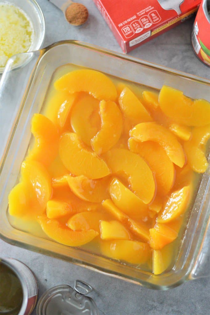 A baking dish filled with peaches with cake mix, cinnamon and melted butter in background