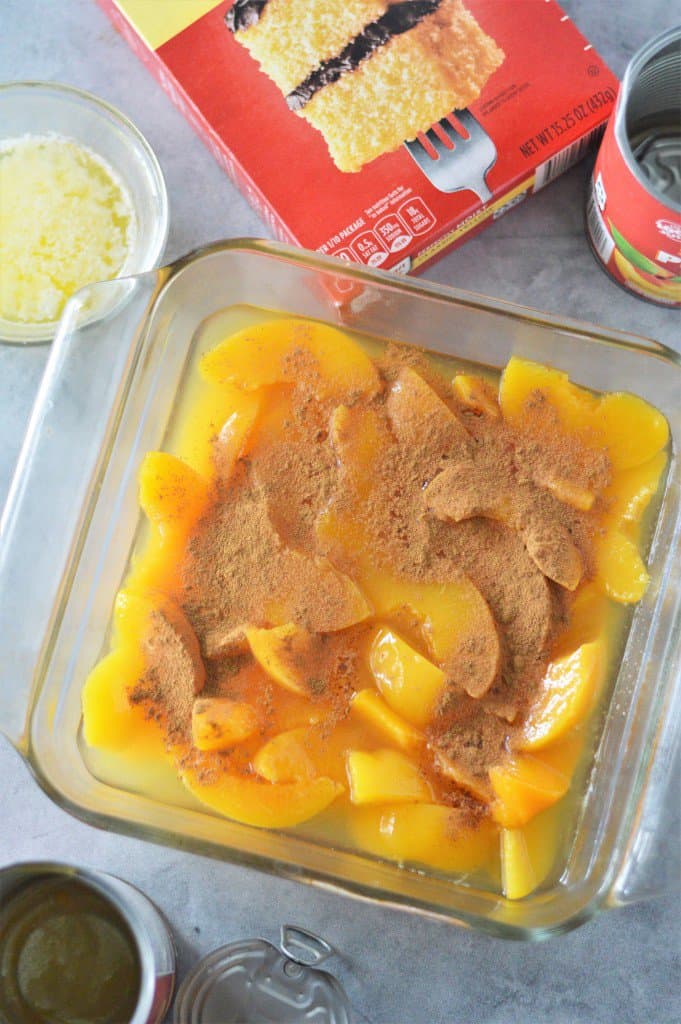 cinnamon on top of canned peaches in a baking dish with cake mix and melted butter in background