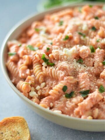 A bowl of creamy blended cottage cheese tomato pasta garnished with herbs and grated cheese, served with a slice of garlic bread on the side.