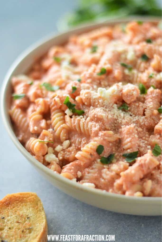 A bowl of creamy blended cottage cheese tomato pasta garnished with herbs and grated cheese, served with a slice of garlic bread on the side.