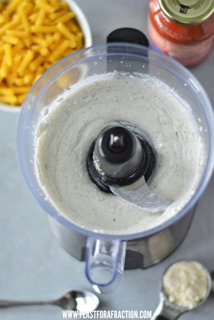 Food processor containing blended cottage cheese, with ingredients in the background.