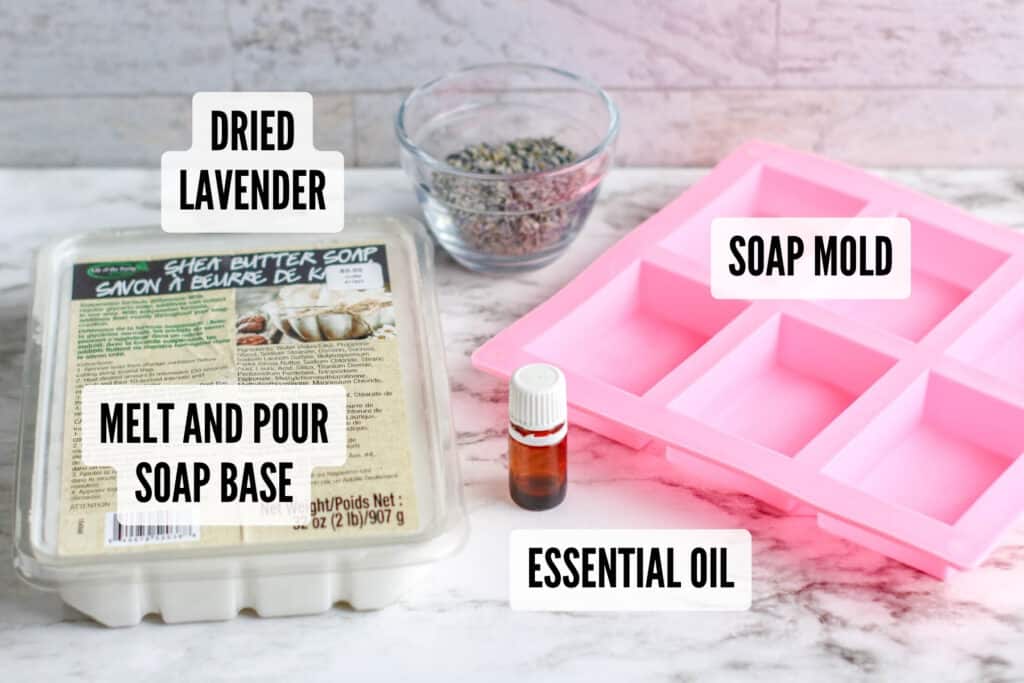 Soap-making supplies on a table including melt and pour soap base, essential oil, dried lavender, and a pink soap mold.