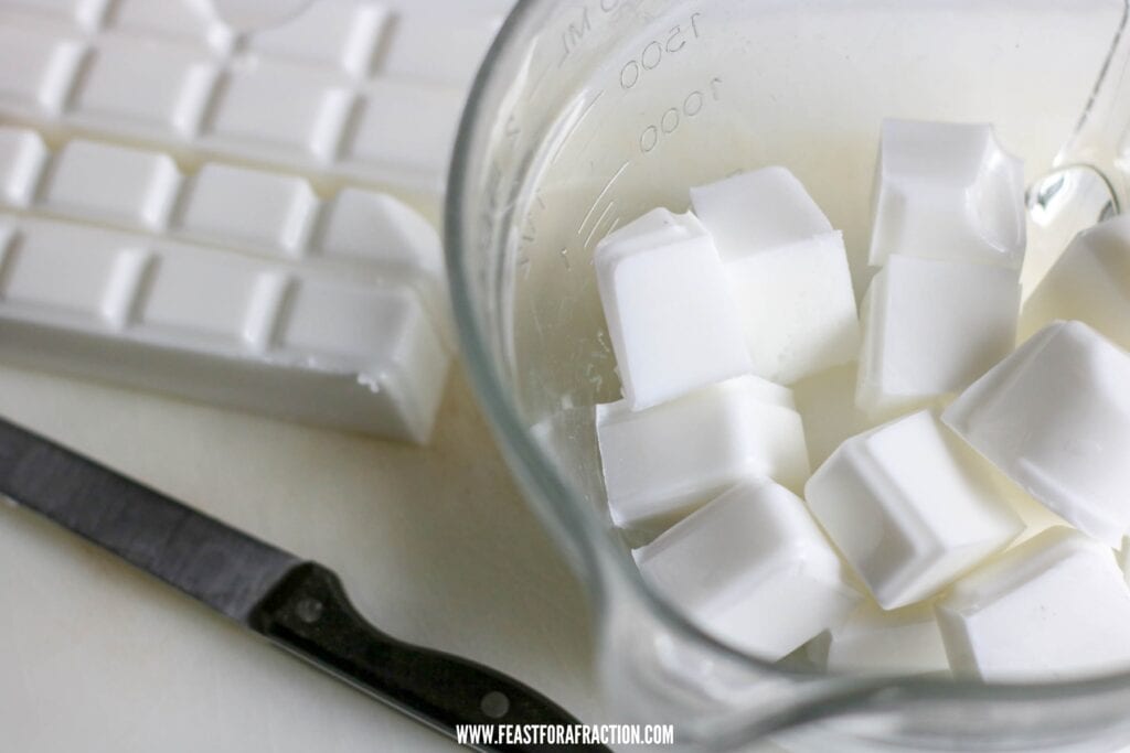 Glass measuring cup with cubed white substances next to a knife and an empty ice cube tray on a white surface.