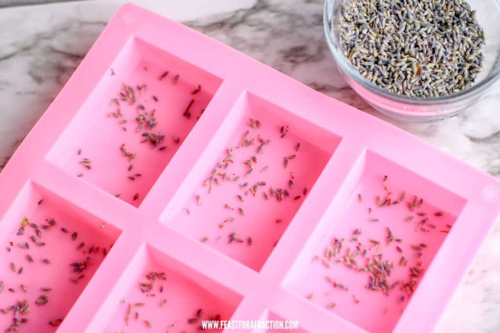 Pink silicone mold filled with lavender-infused soap mixture beside a bowl of lavender buds on a marble surface.
