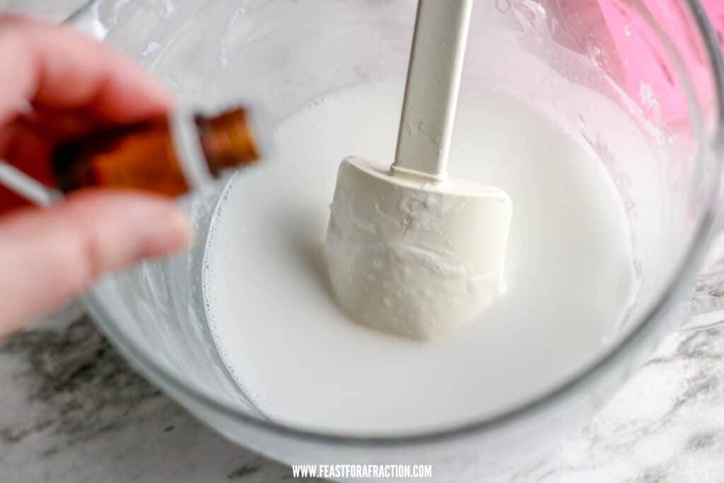 A person dipping a chocolate bar into a glass bowl filled with white icing or cream, using a fork to hold the bar.