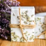 Handmade soap bars with lavender inclusions, tied with twine, displayed next to purple flowers on a wooden surface.