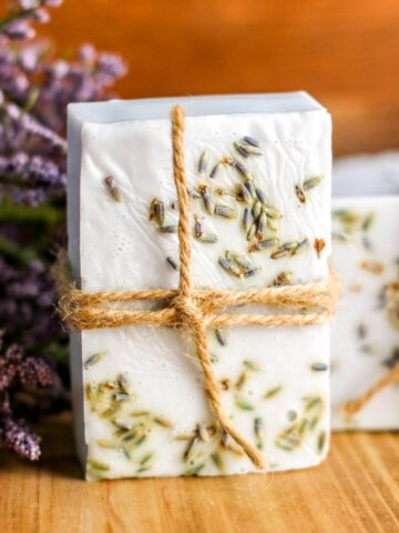 A handmade soap bar with dried lavender and twine, displayed on a wooden surface with lavender sprigs in the background.