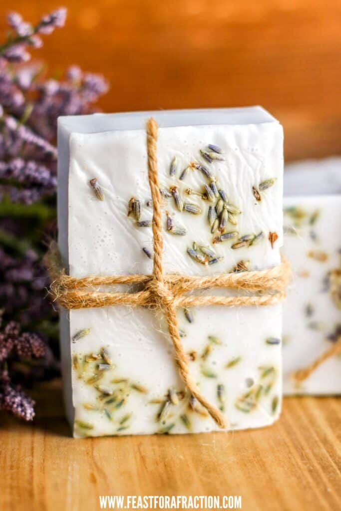 A handmade soap bar with dried lavender and twine, displayed on a wooden surface with lavender sprigs in the background.